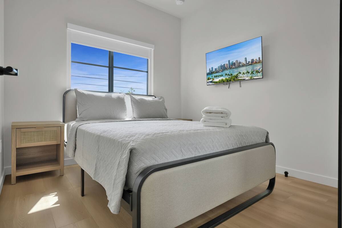 A modern bedroom with a bed, a nightstand, a window with a cityscape view, and a wall-mounted TV.
