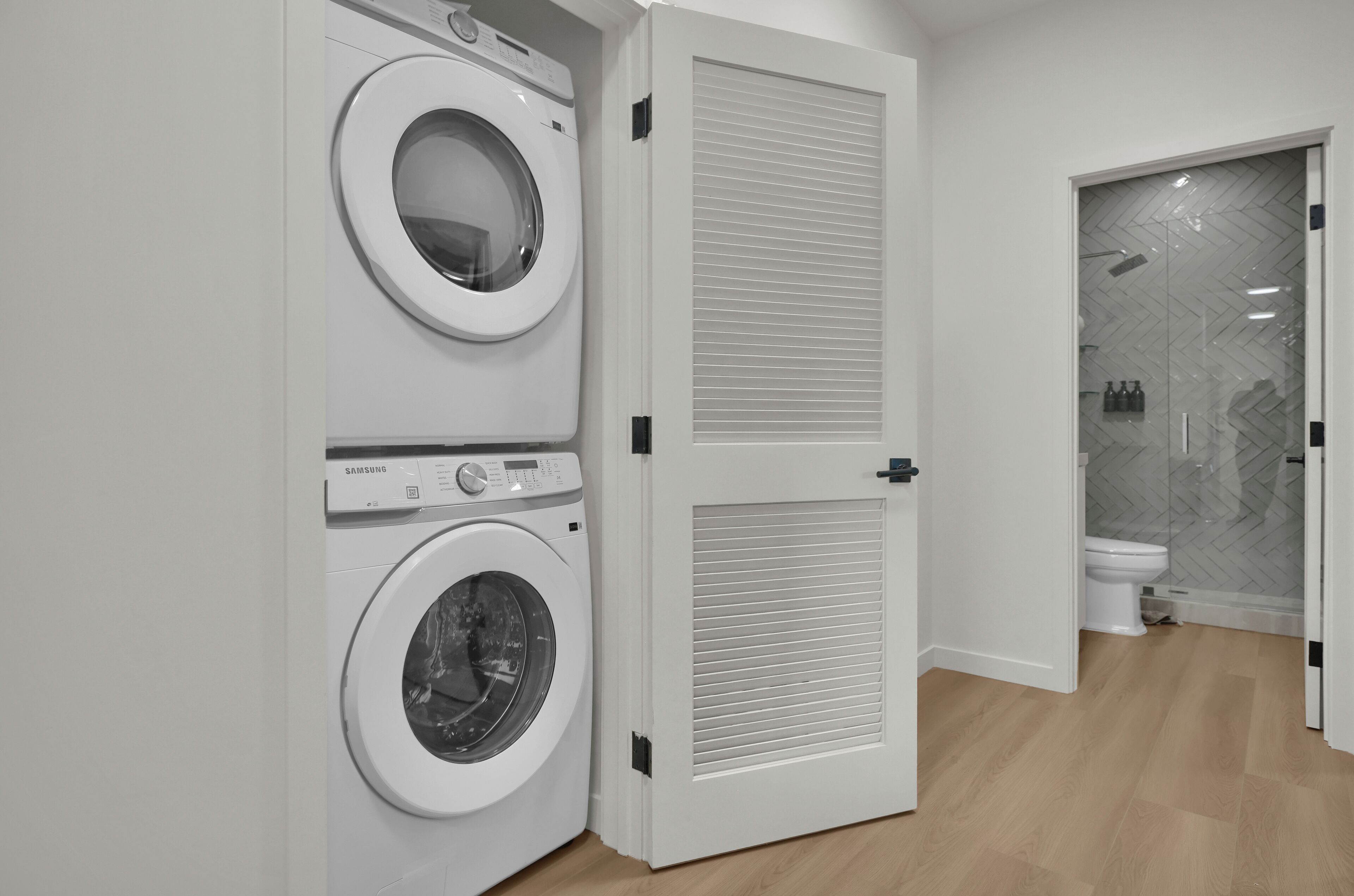 Uma lavanderia com duas máquinas de lavar brancas, uma porta fechada com veneziana e uma área de duche com porta de vidro.
