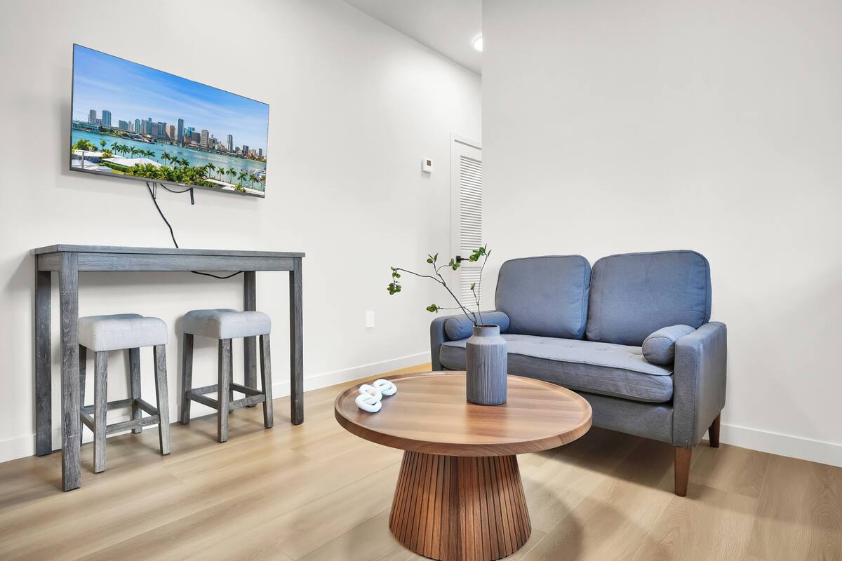 A modern living room with a grey sofa, two bar stools, a round wooden coffee table, and a cityscape painting on the wall.