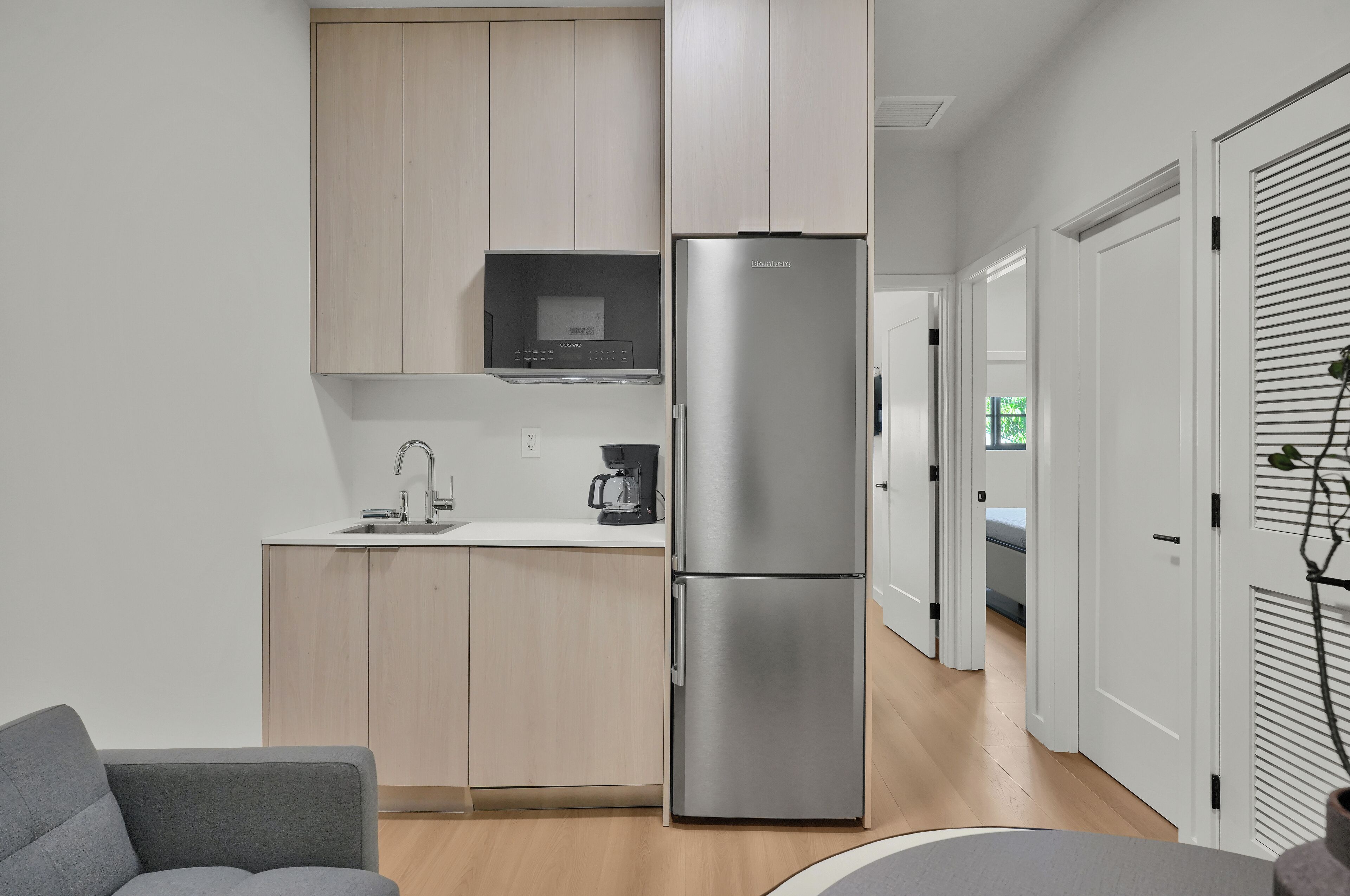 A compact kitchen with a built-in microwave, a stainless steel refrigerator, a coffee maker, and a grey sofa.