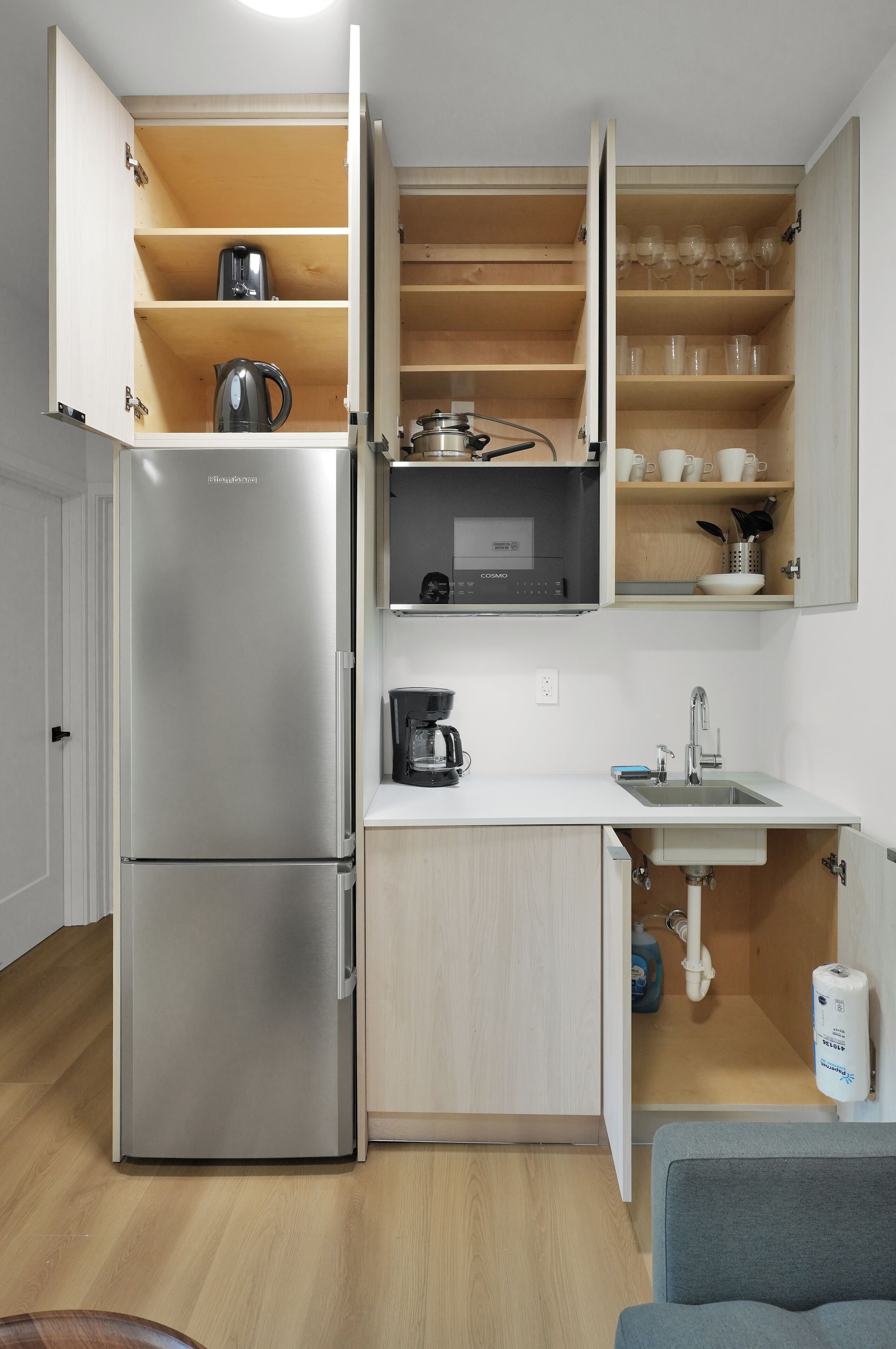 Cocina compacta con refrigerador integrado, cafetera y estantes abiertos con utensilios de cocina.
