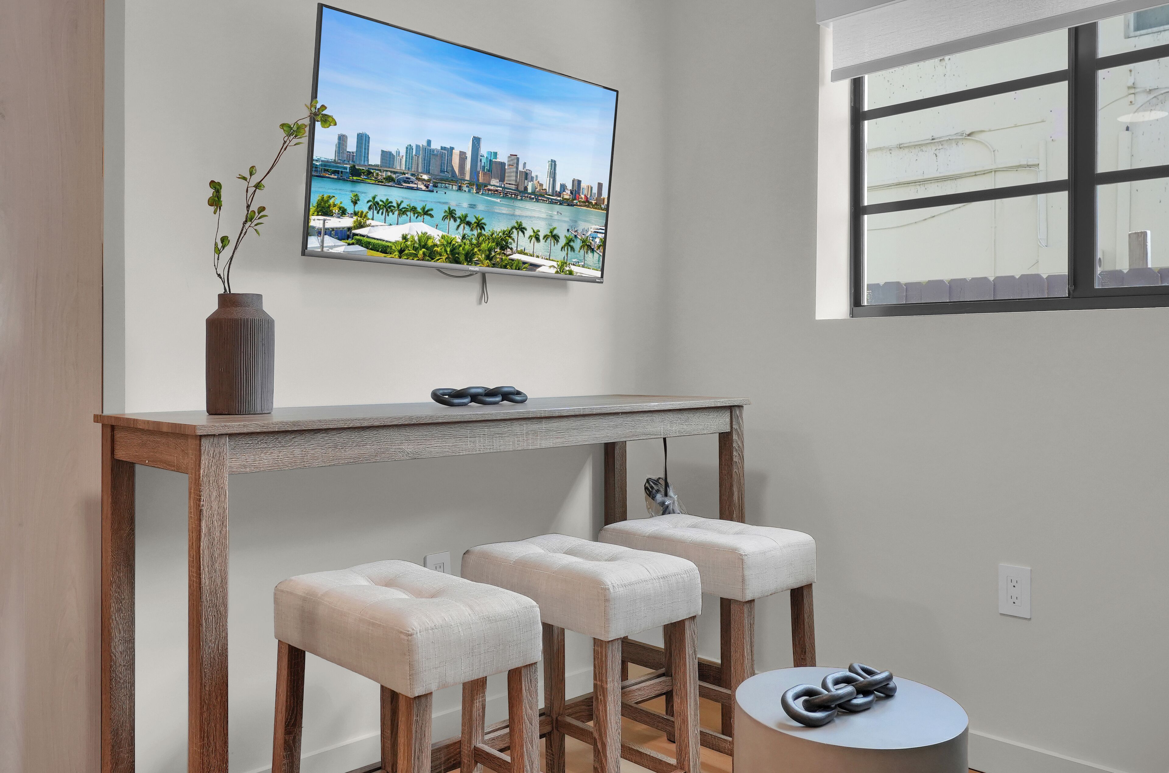 A modern living room with a wooden coffee table, three cushioned bar stools, a flat-screen TV mounted on the wall, and a window with blinds.