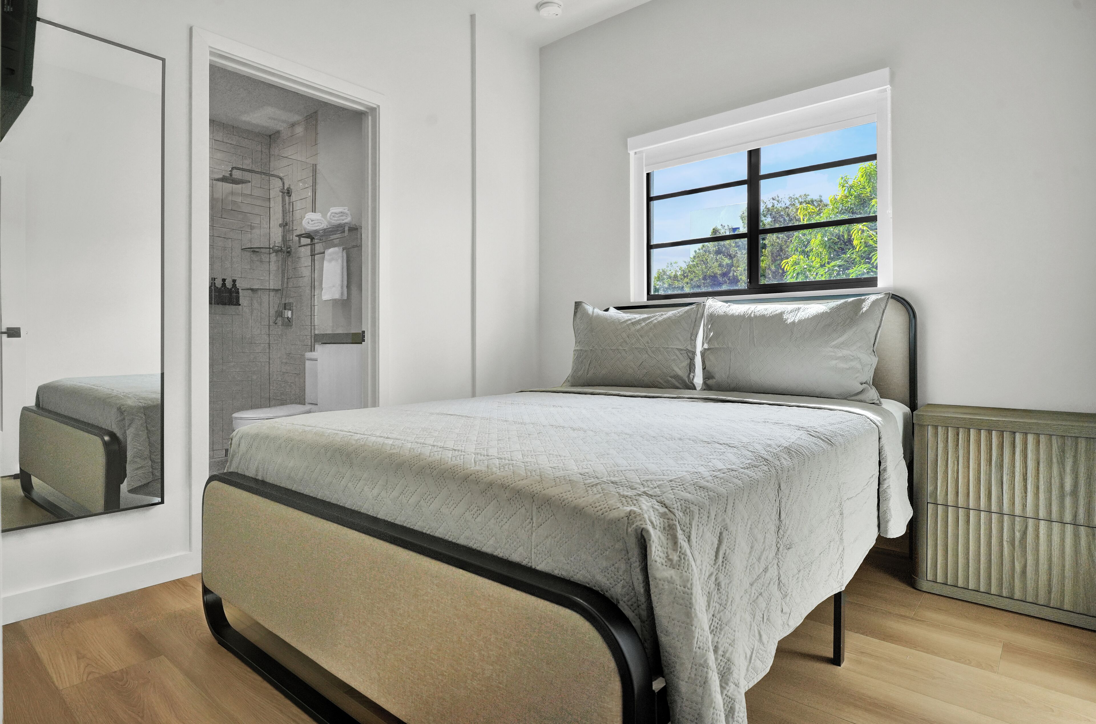 A bedroom with a bed, a window with a view, a radiator, and a bathroom with a shower and toilet.