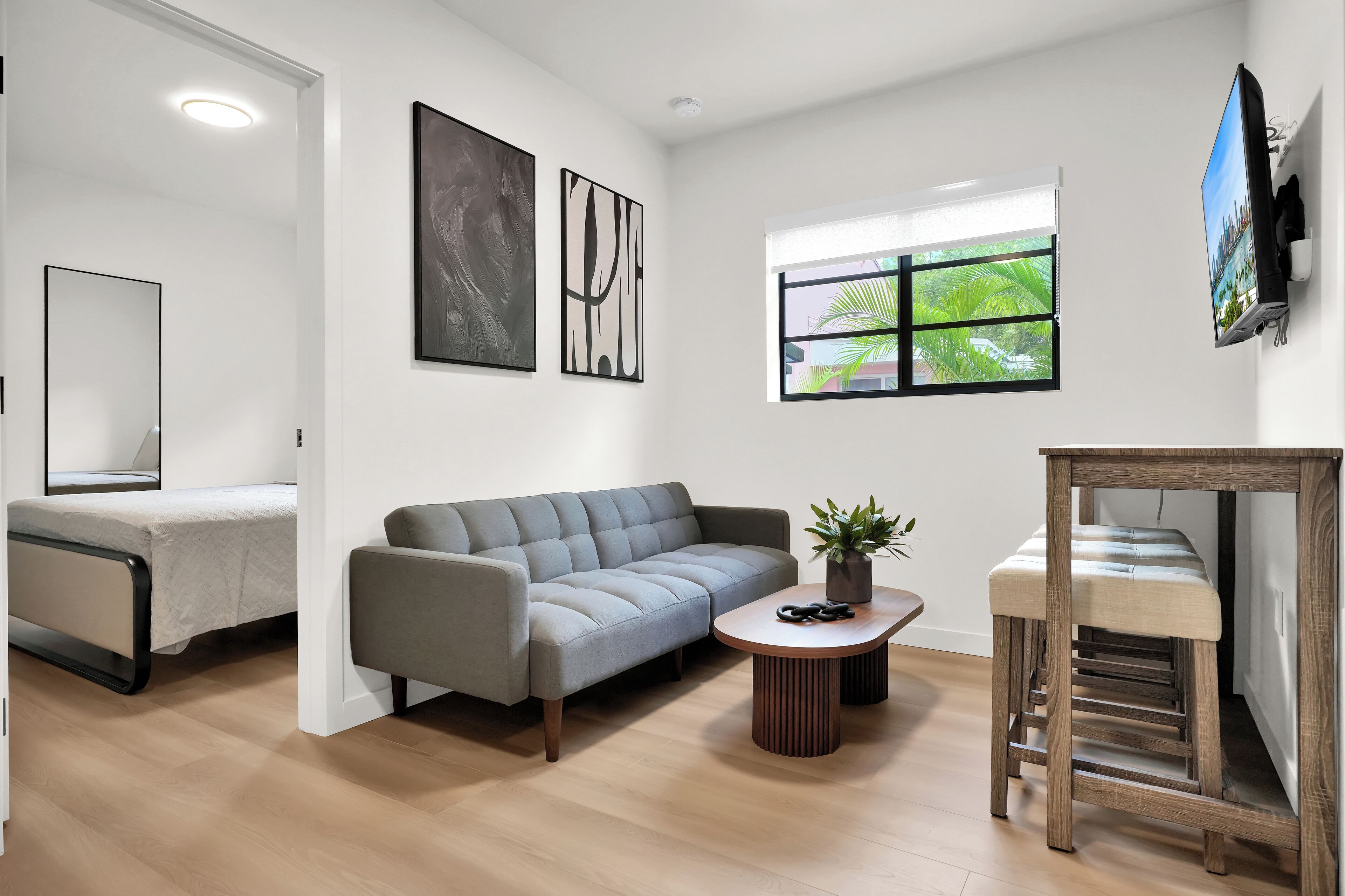 Uma sala de estar moderna com um sofá cinzento, uma mesinha redonda com uma planta, um banquinho de madeira e uma TV fixada na parede.