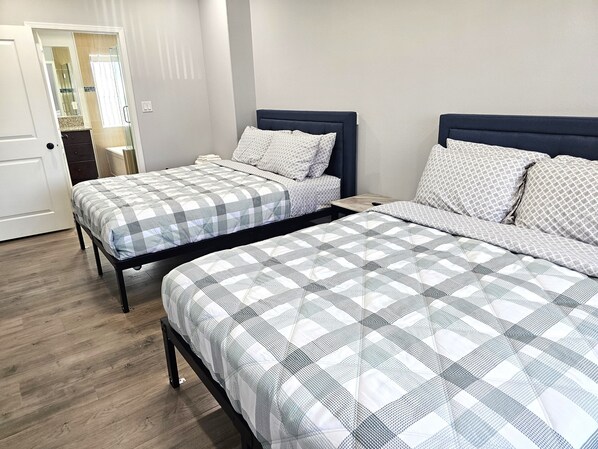 Une chambre avec deux lits, chacun recouvert d’une courtepointe à carreaux et agrémenté d’oreillers assortis. Le sol est en bois et, au fond, on aperçoit une porte blanche.