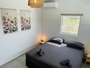 A bedroom with a bed, two wall-mounted artworks, a bedside table with a lamp, and a window with a view of a garden.