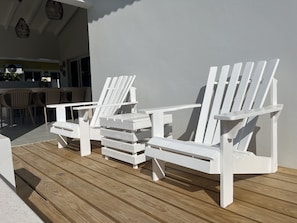 Two white Adirondack chairs on a wooden deck with a white wall in the background.