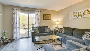 En moderne stue med to grå sofaer, et glasbord og et spisebord med stole. Der er en balkon med udsigt til træer og en vægkunst med et cykeltema.