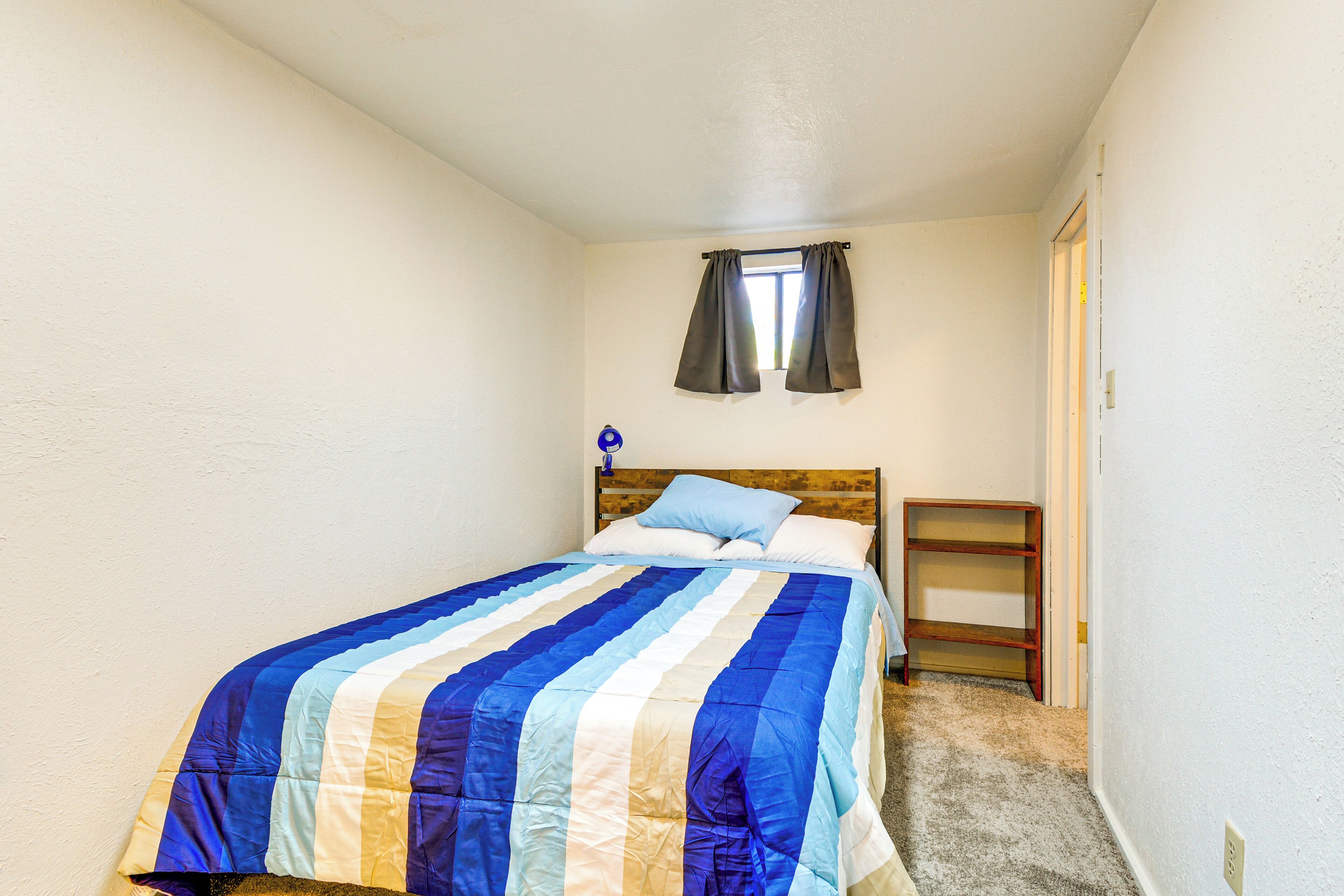 Um quarto com cama, janela com cortinas, mesa de cabeceira e prateleira de madeira.