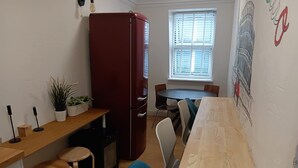 A compact kitchen with a refrigerator, a dining table with chairs, a small countertop with stools, and a window with blinds.