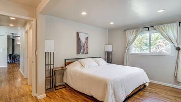 Une chambre avec un grand lit, un plancher en bois, une table de chevet, une commode, une fenêtre avec des rideaux et un tableau accroché au mur.