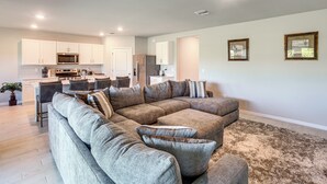 A modern living room with a sectional sofa, a dining area with a kitchen in the background, and a large window providing natural light. - Family Home w/ Patio & Canal Views in Cape Coral! (Cape Coral)