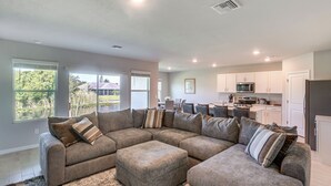 A spacious living room with a sectional sofa and ottoman, a kitchen area with modern appliances, and large windows offering a view of greenery. - Family Home w/ Patio & Canal Views in Cape Coral! (Cape Coral)