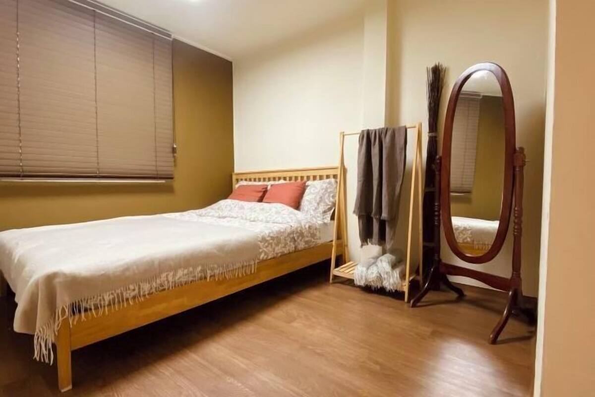 A single bed with a wooden frame, a bedside table, a mirror, and a window with blinds.