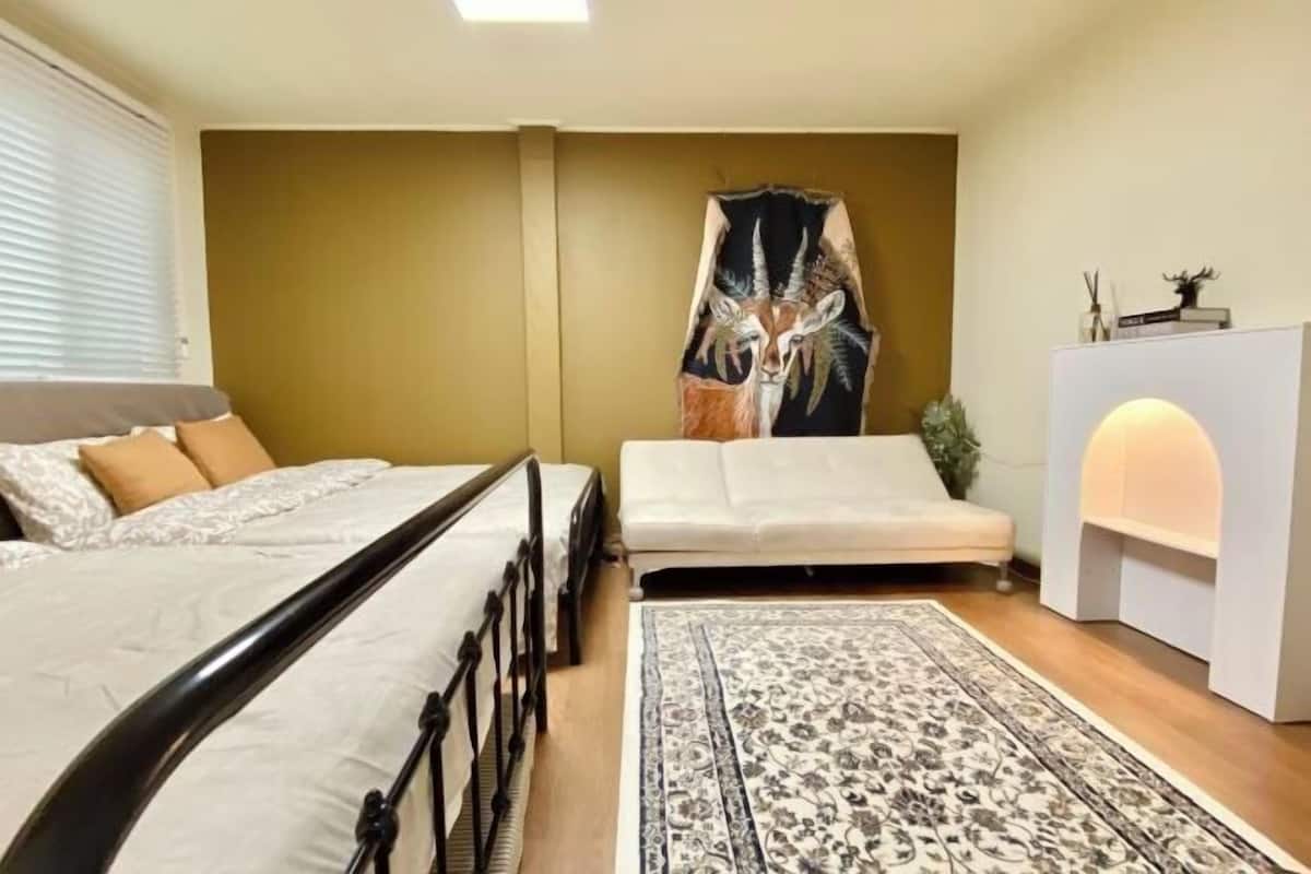 A bedroom with a bed, a sofa, a rug, a wall tapestry, and a bedside table with a lamp.