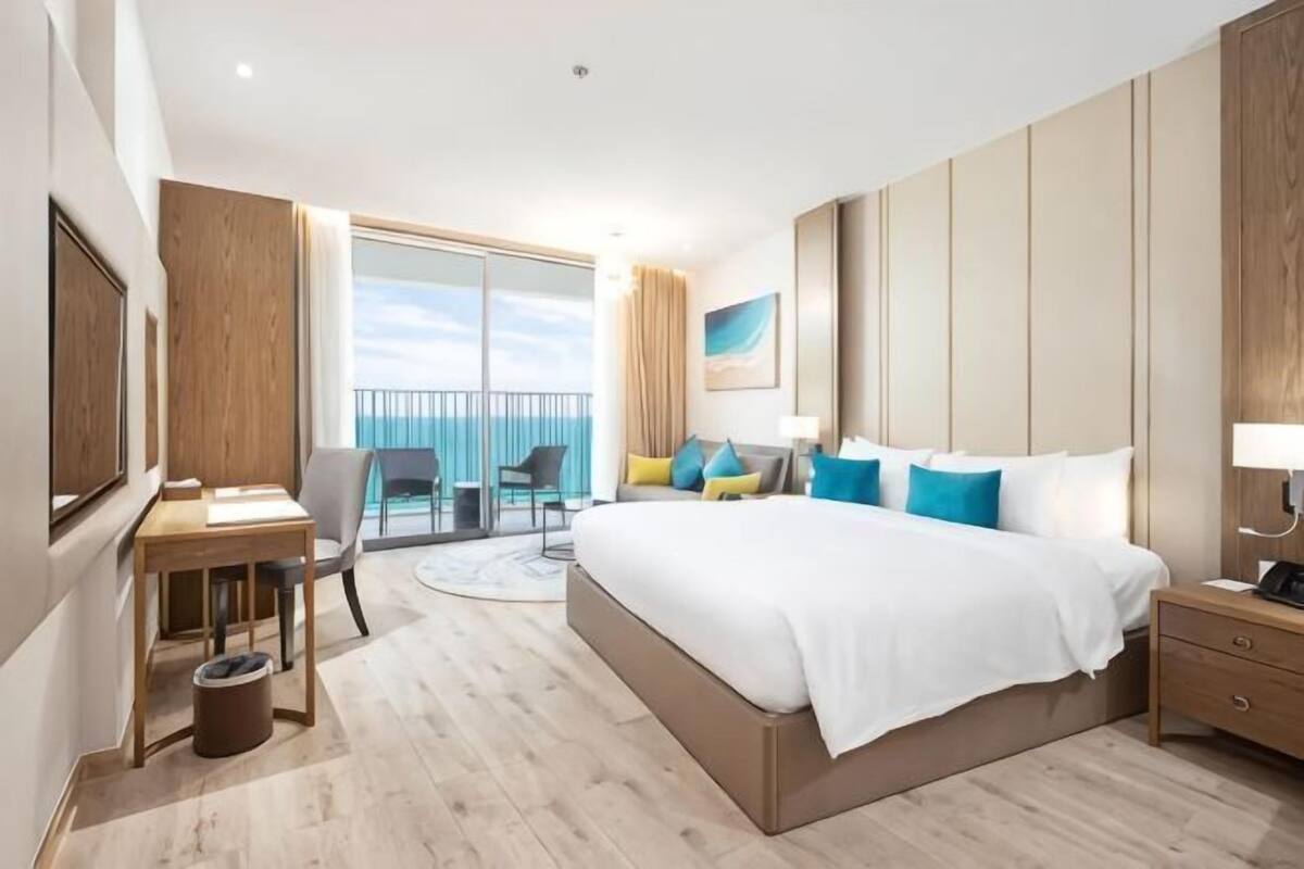 Una camera d'albergo con un letto grande, una scrivania, una sedia, un tavolino, un comodino e un balcone vista mare.