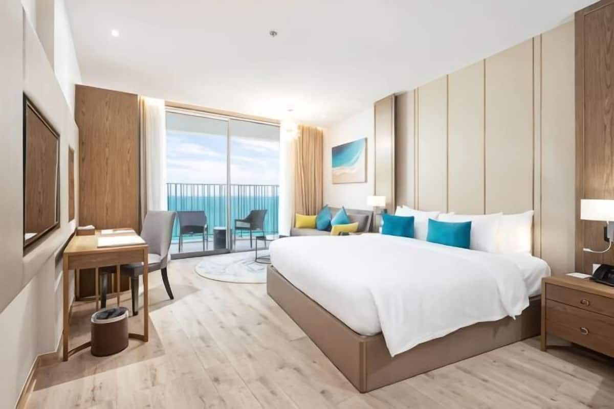 Ein Hotelzimmer mit einem großen Bett, einem Schreibtisch, einem Sessel, einem kleinen Tisch, einem Nachttisch und einem Balkon mit Meerblick.