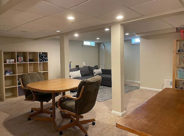 A room with a wooden table and chairs, a bookshelf, a gray sofa, and a carpeted floor.