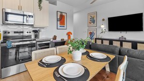 Una cucina moderna con elettrodomestici in acciaio inossidabile, una zona pranzo con tavolo in legno apparecchiata per quattro persone e un ampio televisore a schermo piatto montato a parete.