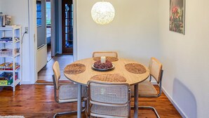 A dining area with a round wooden table, four wicker chairs, a hanging light fixture, and a bookshelf with various items.