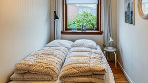 A narrow room with a bed, a small table, a lamp, and a window with a view of greenery and a building outside.