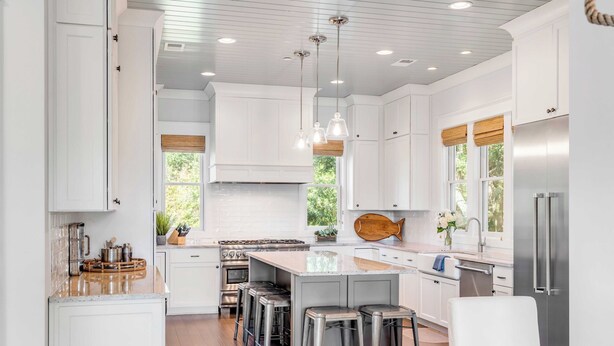 Una cocina moderna con armarios blancos, una isla central con taburetes, electrodomésticos de acero inoxidable e iluminación colgante.