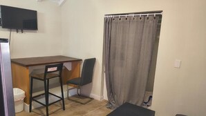 Une petite pièce, de taille modeste, comprenant un bureau, deux chaises, un téléviseur, un rideau gris et un banc.