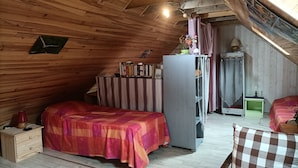 A cozy attic bedroom with a bed, a dresser, a table with books, a refrigerator, and a window with curtains.