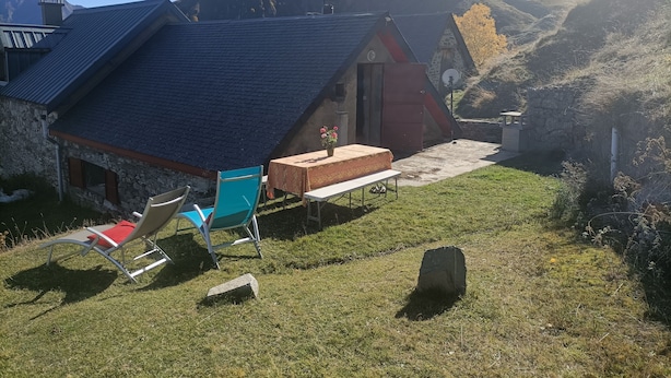 A small stone building with a sloped roof, a patio with a table and chairs, and a view of mountains in the background.