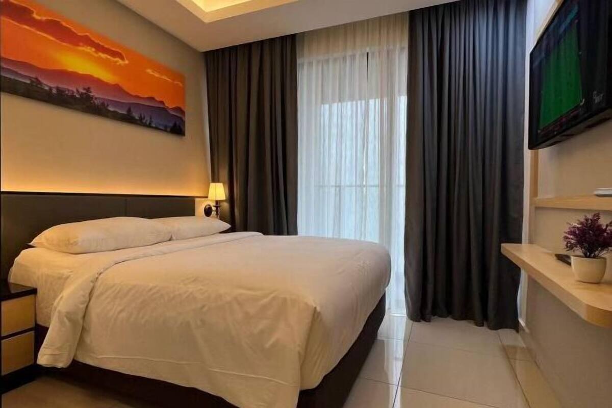 Ein Hotelzimmer mit einem großen Bett, einem Nachttisch mit Lampe, einem Gemälde an der Wand, einem an der Wand befestigten Fernseher und einem Fenster mit Vorhängen.