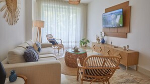 Een moderne woonkamer met een bank, twee rieten fauteuils, een glazen salontafel, een houten kast en een televisie die aan de muur is bevestigd.