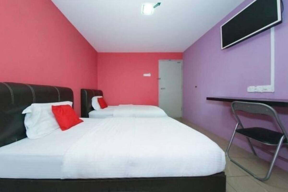 A room with three beds, each with white bedding and red pillows, pink walls, and a television on the wall.