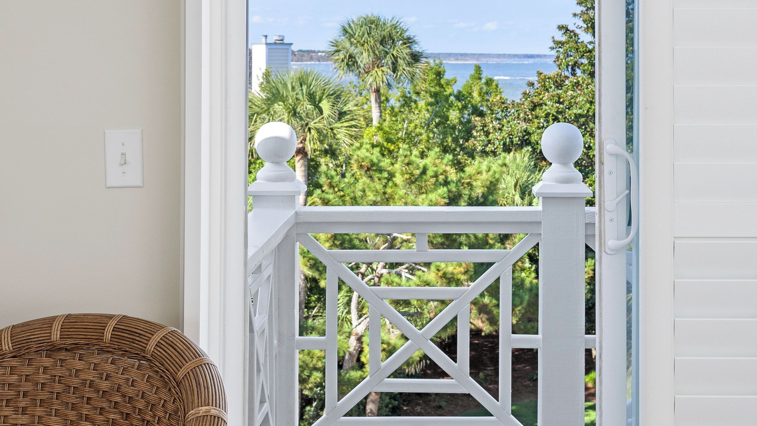A balcony with a white railing, a wicker chair, and a view of a coastal landscape with trees and the ocean.