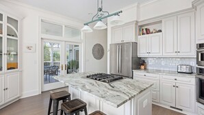 Une cuisine moderne avec des meubles blancs, un îlot en marbre, des appareils en acier inoxydable et un coin repas doté d’une porte en verre donnant sur l’extérieur.