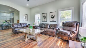 Amplia sala de estar con sofá de cuero, una mesa de centro y un sillón de cuero. Hay cuadros enmarcados en la pared, un ventilador de techo y vistas a la cocina a través de una puerta abierta.