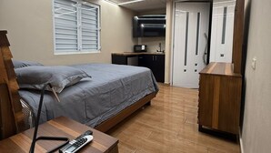 A modern bedroom with a bed, bedside table, and a desk with a chair. There is a window with blinds, a microwave, and a TV mounted on the wall. The room has a wooden floor and a door leading to another room. - Caleno Boricua apartment. Modern luxury apartment 5 minutes from the beach (Quebradillas)