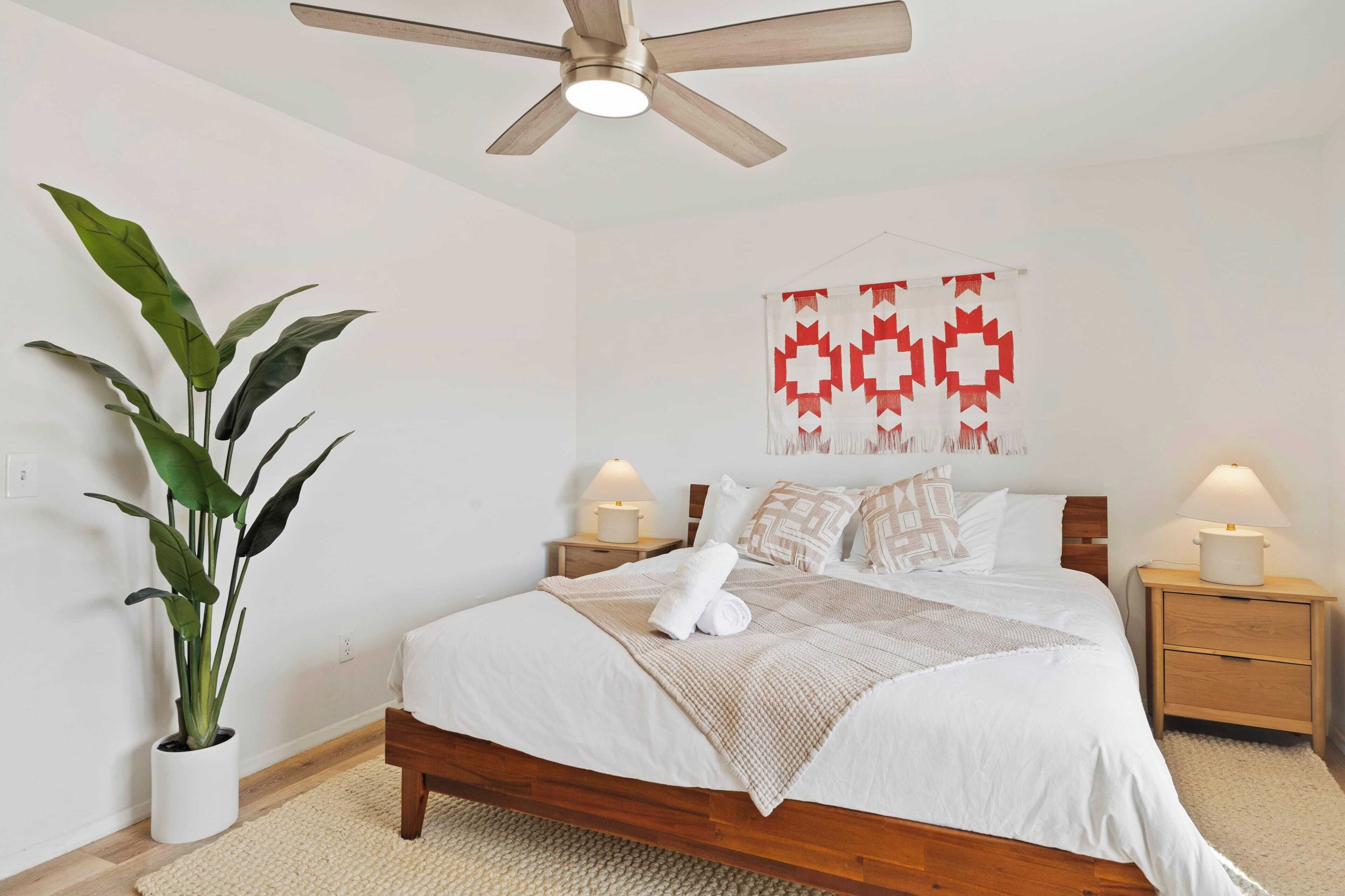 Un lit bien fait, avec du linge de lit blanc et un plaid beige, une plante en pot, un ventilateur de plafond et une décoration murale à motifs géométriques.