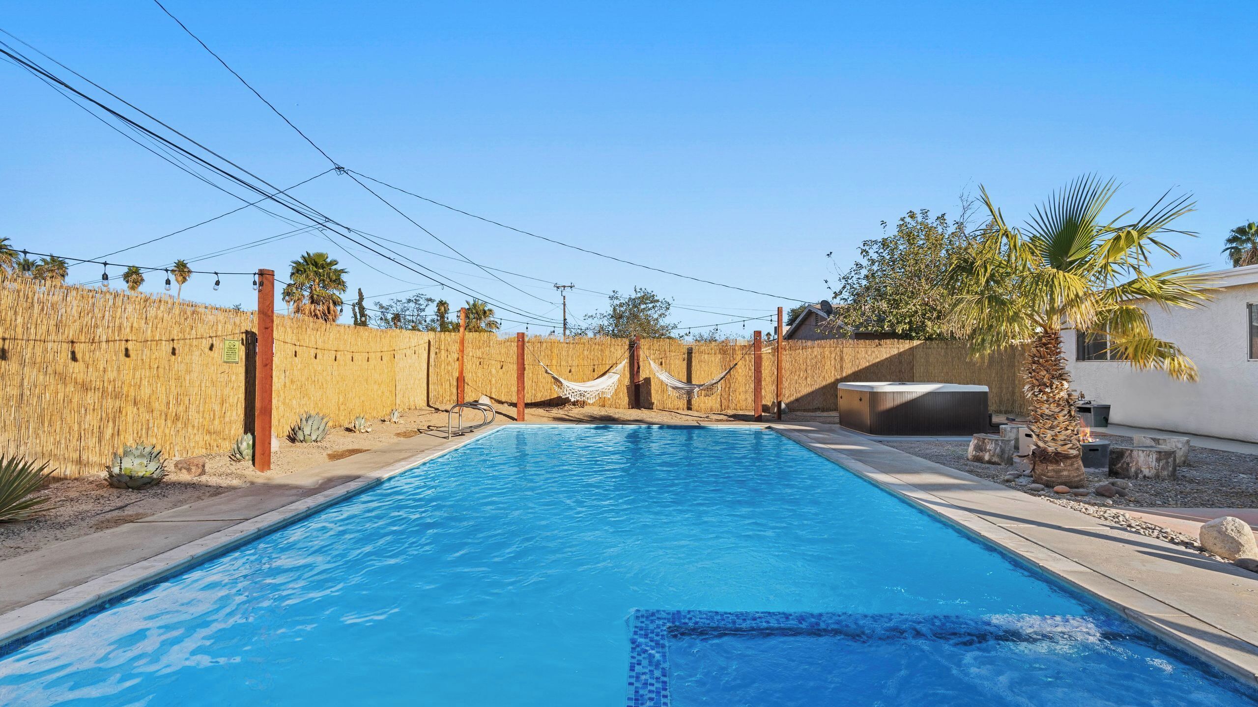 A rectangular swimming pool with a tiled edge, surrounded by a wooden fence, a hammock, and a hot tub.