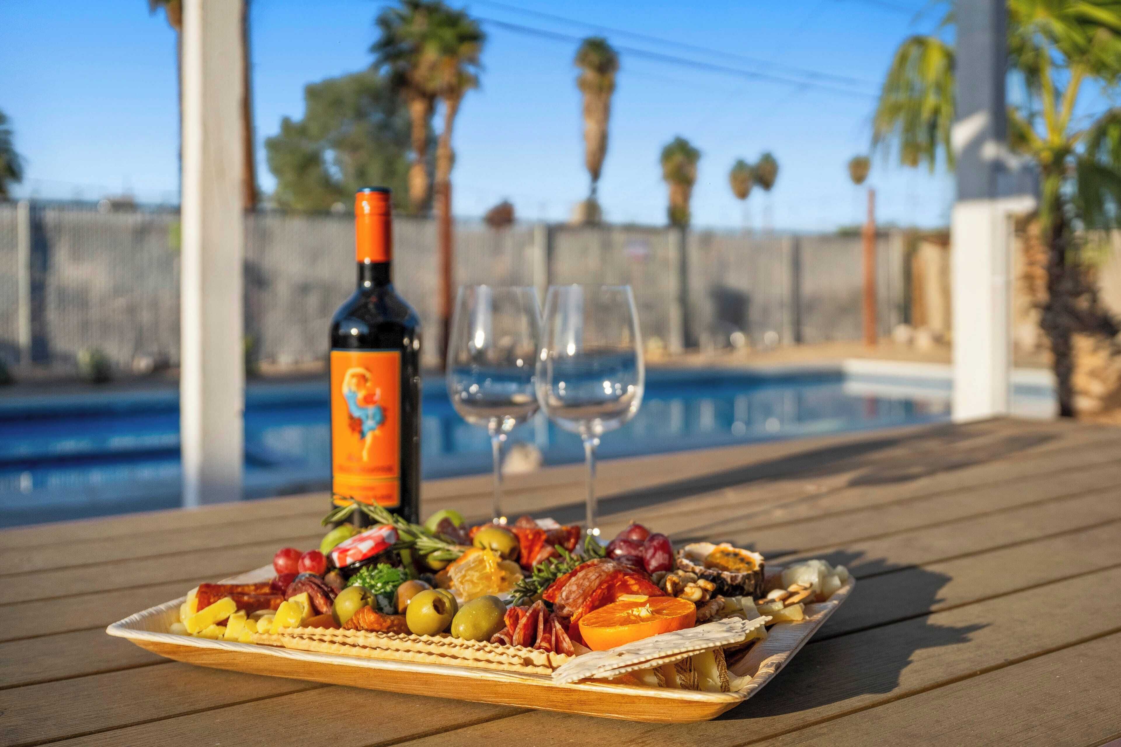 Une planche de charcuterie avec différentes sortes de viandes, de fromages et de fruits, posée sur une table en bois au bord d’une piscine.