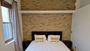 A bedroom with a brick wall, a large bed with white bedding, two bedside tables, and a window with curtains. - Downtown Depot - Unit 5 (Fargo)