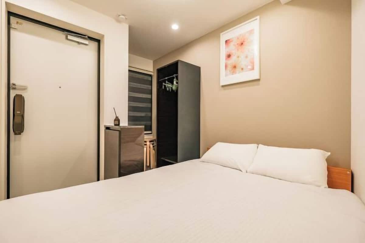 A hotel room with a bed, a wardrobe, a desk with a chair, and a framed picture on the wall.