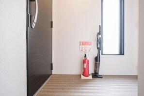 Una habitación con suelo de madera, una puerta con manija, una ventana y un extintor de incendios sujeto a la pared.