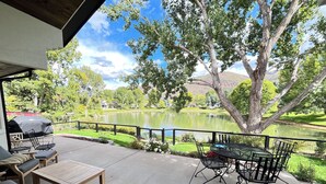 Eine Terrasse im Freien mit Tisch und Stühlen, einem Grill und Blick auf einen See und umliegendes Grün.