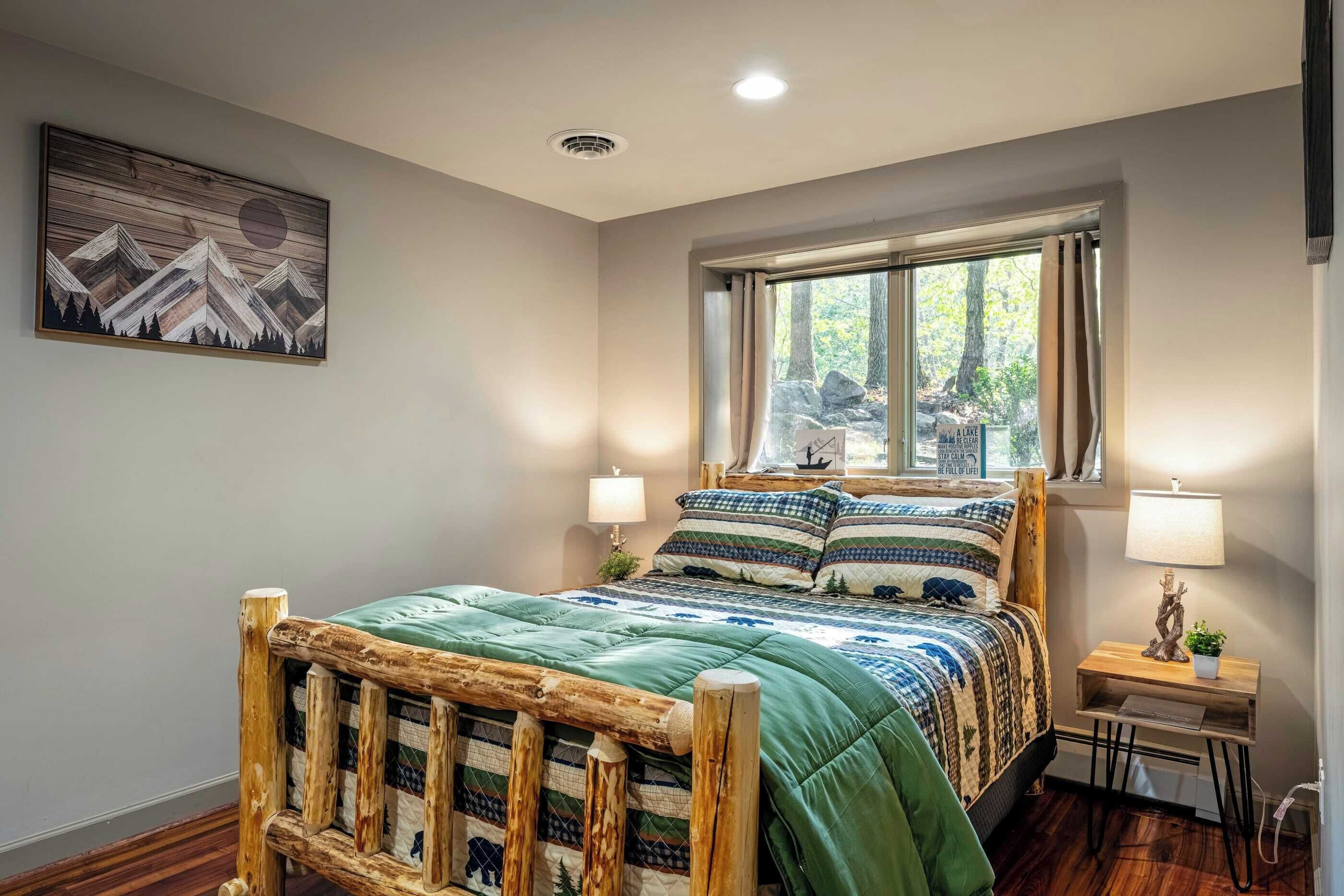 A bedroom with a wooden bed frame, a green bedspread, two bedside lamps, a nightstand with a plant, and a window with a view of trees.