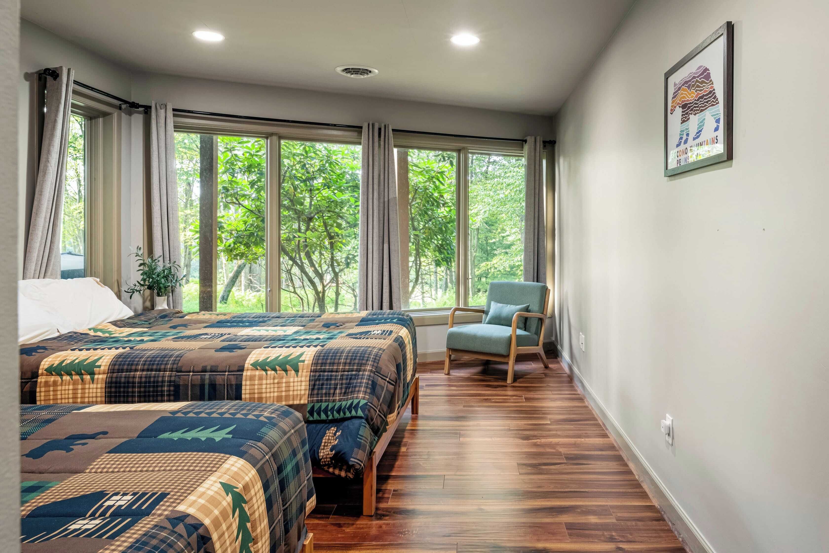 A bedroom with two beds, a chair, and large windows with a view of trees.