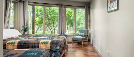 A bedroom with two beds, a chair, and large windows with a view of trees.