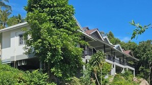 Ein modernes, zweistöckiges Haus mit Balkon, umgeben von üppiger Vegetation und Palmen.