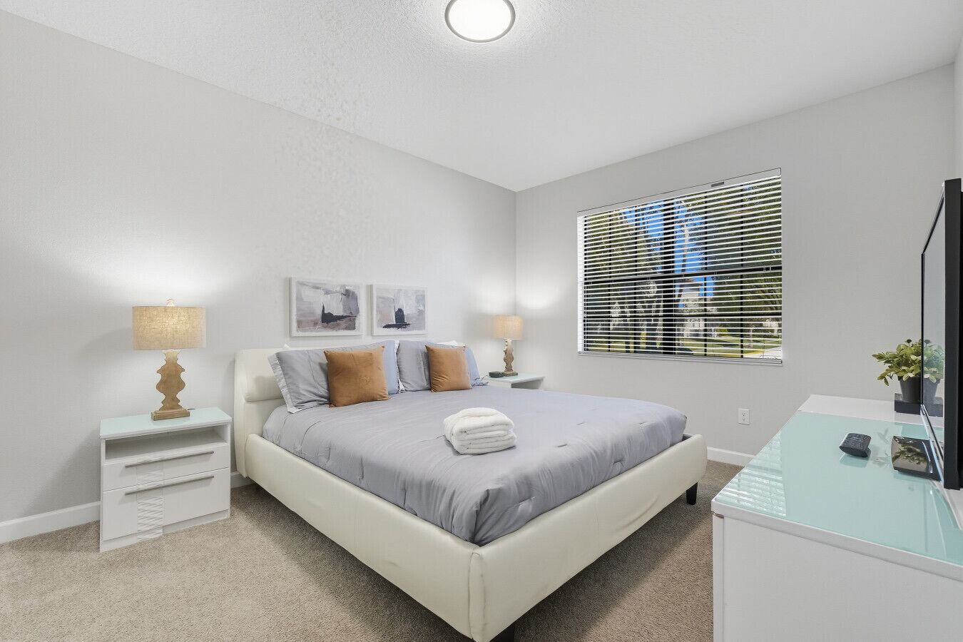 Une chambre à coucher comprenant un lit, des tables de chevet, une lampe, une fenêtre avec des stores et un petit bureau agrémenté d’une plante.
