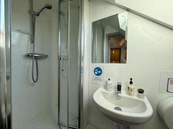 Un baño con ducha de vidrio, lavamanos con gel antibacterial y espejo.