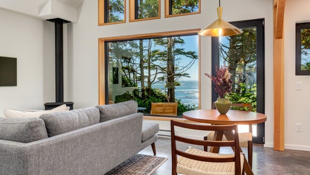 A modern living room with a gray sofa, a wooden dining table with chairs, and large windows offering a view of trees and the ocean. - Fogged Inn (Ucluelet)
